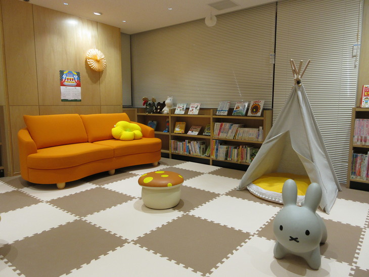 ベージュとホワイトの格子のカーペットが敷き詰められた部屋にオレンジ色のソファー、キノコやウサギ型の椅子、小型のテントや本棚が設置されている写真